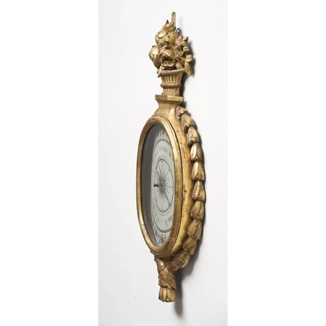 18th century French giltwood oval barometer by Torre, the crest a carved basket of flowers, the sides with cascading...
