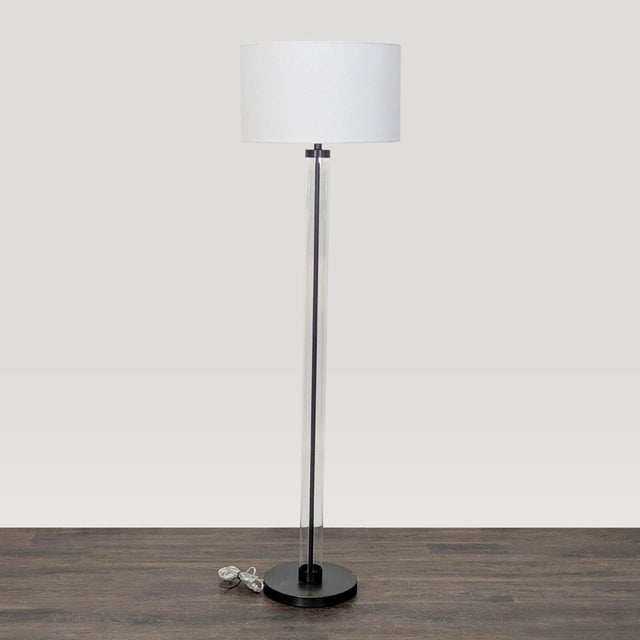 This Arhaus floor lamp features a sleek clear glass cylinder base with a sturdy black foundation and a simple white drum...