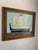 This seascape pairs a robin’s egg blue sky with an avocado green foreground and black vessel, softened by off-white sails....