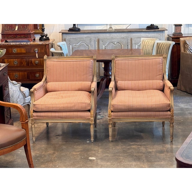 A pair of carved and giltwood bergere armchairs upholstered in a rose and cream striped fabric, with loose seat and back...