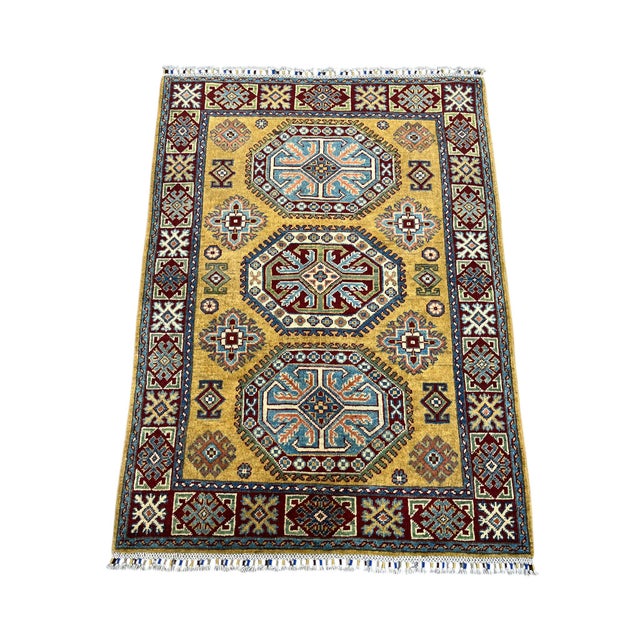 Three bold medallions run down the warm gold field of this Kazak rug. Burgundy and light blue motifs add contrast, while a...