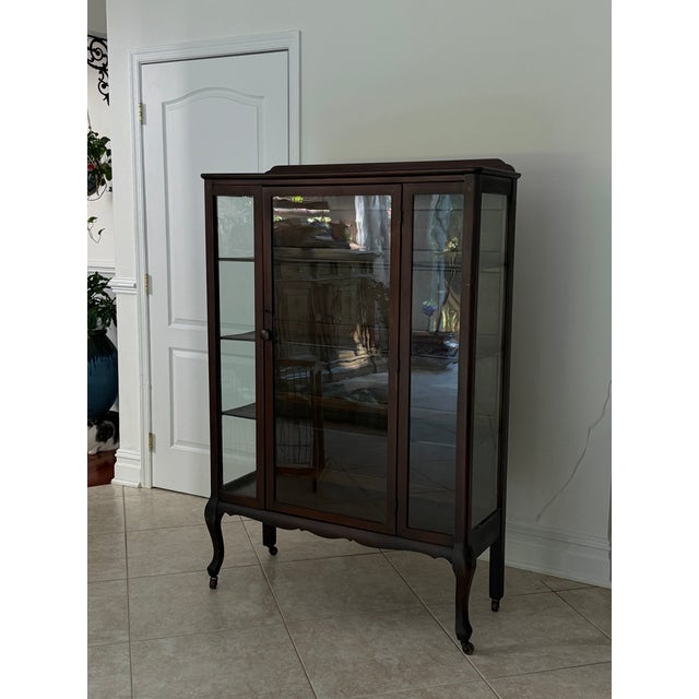 Elegant antique glass display cabinet featuring a rich, dark mahogany wood frame, graceful cabriole-style legs on casters,...