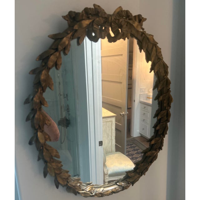 A refined Neoclassical-style mirror framed by a sculptural laurel wreath, carved in relief and finished in a warm, softly...