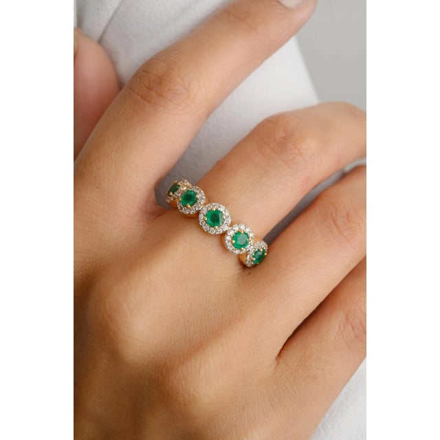 This elegant wedding band features five natural round-cut emeralds, each encircled by a dazzling halo of diamonds. The...