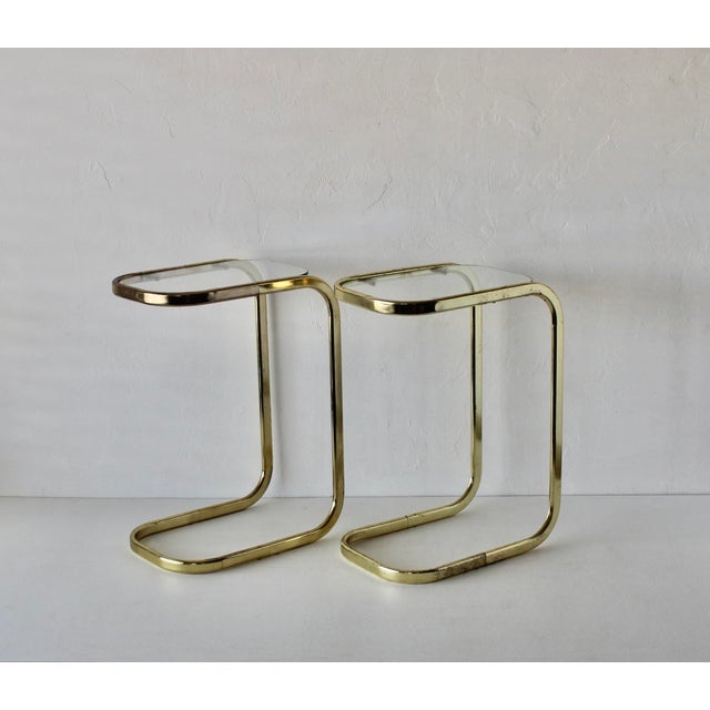 A pair of Mid-Century Modern brass-finished and glass cantilever side tables, often attributed to the design of Milo...