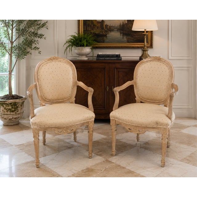 Elegant pair of vintage Louis XVI style armchairs featuring carved wood frames with a softly distressed, warm cream...