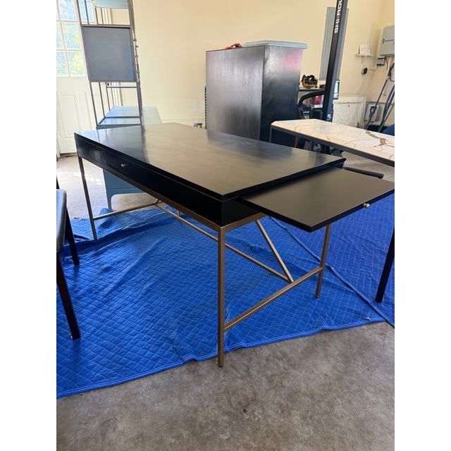 Elegant black writing desk featuring a sleek, expansive work surface paired with a slender brass-finish metal base. The...