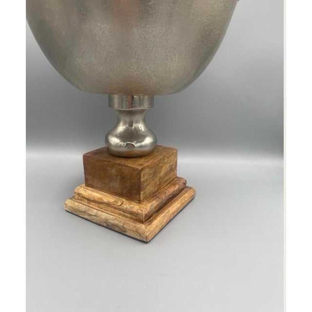 Elegant silver-tone metal pedestal bowl displayed on a substantial carved wood base. The combination of the cool-toned...