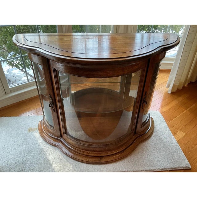A graceful demilune console curio with a shaped serpentine top, curved glass on the front and sides, and a molded plinth...