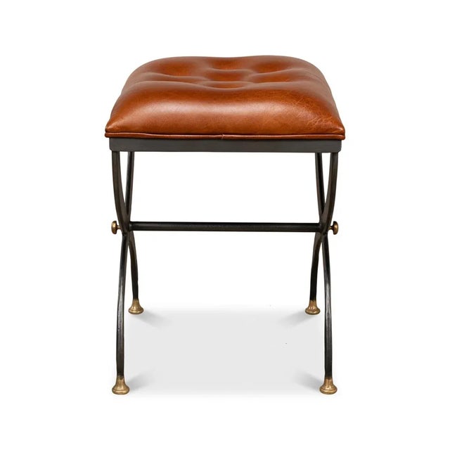 A Modern curule stool with a button tufted leather cushion seat above a pewter-finished iron curule form base with gilded...