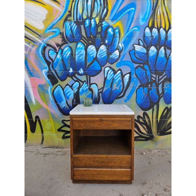 A beautifully composed vintage mid-century nightstand featuring warm oak grain contrasted by a polished marble top surface...
