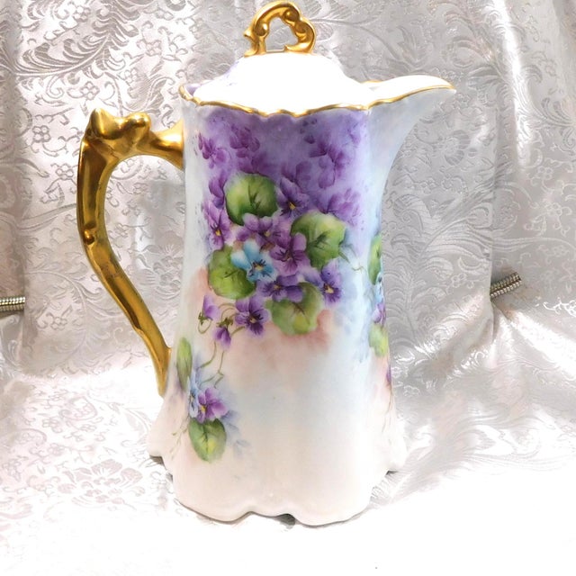 This is a white chocolate pot with a purple flower design with a single blue flower and some pink. It has a gold painted...