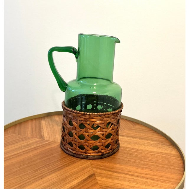 Beautiful vintage green blown glass pitcher featuring a warm woven wicker basket wrap. The rich emerald tone, hand-applied...