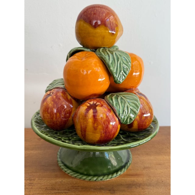 A colorful ceramic fruit topiary of peaches and oranges stacked on a deep green footed pedestal. Glossy glazed fruit with...