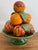 A colorful ceramic fruit topiary of peaches and oranges stacked on a deep green footed pedestal. Glossy glazed fruit with...