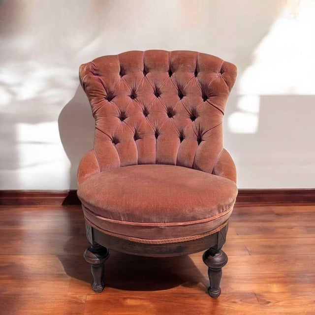 Victorian style tufted slipper chair with pink velvet upholstery and a carved wood base. The chair has a rounded fan-like...