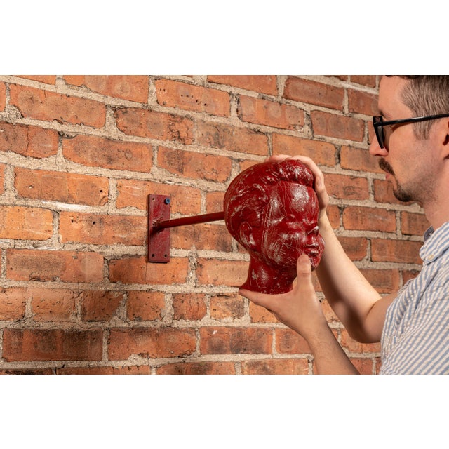 Painted metal display head mounted on a metal wall bracket, modeled with exaggerated mid-century facial features including...