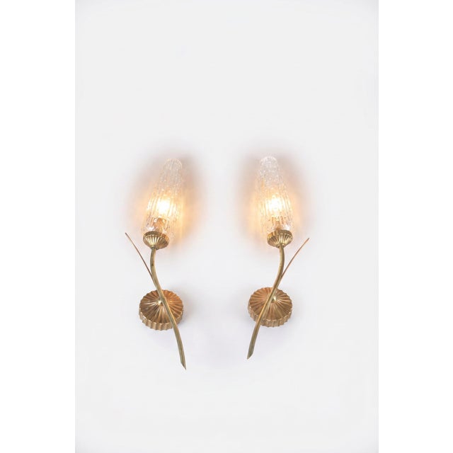 Pair of wall sconces dating to the 1970s. Each has a curved gold-toned metal stem and slender decorative accents that...