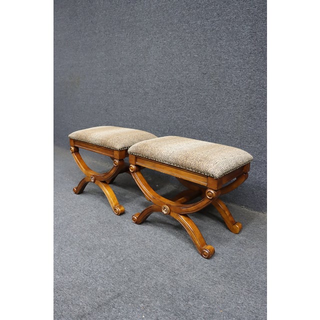 Pair of upholstered stools with curved X-base legs in a warm wood finish. The frames feature round rosette details at the...