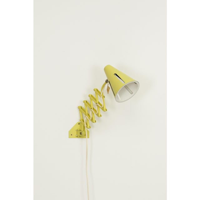 A scissor wall lamp produced by hala zeist in the 1950s. The lamp features a yellow metal shade and an adjustable scissor...