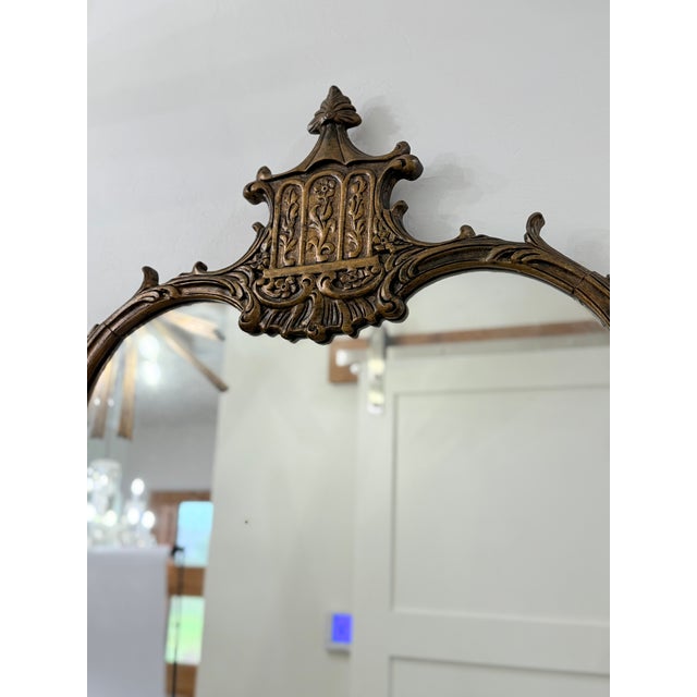 This gilded mirror features an intricately carved wooden frame crowned with a stylized pagoda top. The aged patina lends...