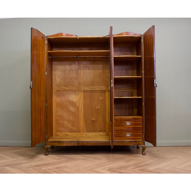 A 1930s Art Deco wardrobe in figured walnut veneer, with three shaped doors and decorative metal handles. Inside are...