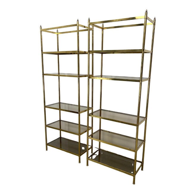 A pair of Billy Baldwin style étagères featuring clean-lined brass frames supporting graduated smoked glass shelves. The...