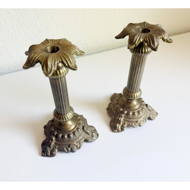 A classic pair of vintage ornate cast brass candlesticks featuring fluted neoclassical columns and stylized palm leaf or...
