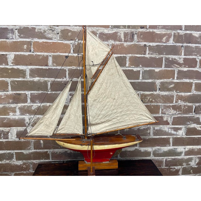 A vintage pond yacht, sloop design with a main mainsail and multiple smaller head sails. The hull is wooden with a cream...