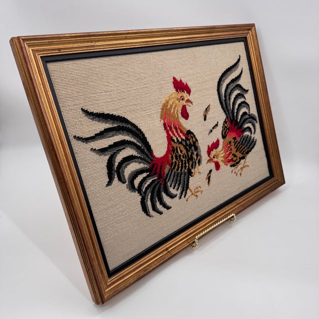 Vintage framed needlepoint featuring a dramatic pair of fighting roosters in rich red, golden ochre, and deep black tones...