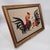 Vintage framed needlepoint featuring a dramatic pair of fighting roosters in rich red, golden ochre, and deep black tones...