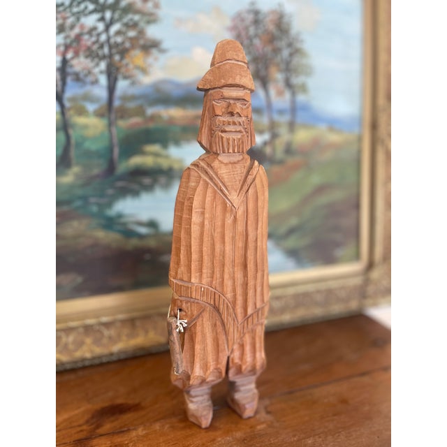 This hand-carved folk art wood sculpture depicts a bearded man wearing a wide-brimmed hat and long cloak, carrying a small...