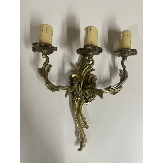 A set of eight gilt bronze Rococo style three-arm wall sconces with scrolling acanthus leaf arms curving outward to...