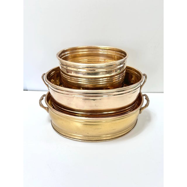 Three vintage oval brass cachepots in graduated sizes, with raised bands and rounded rims. The two larger planters have...
