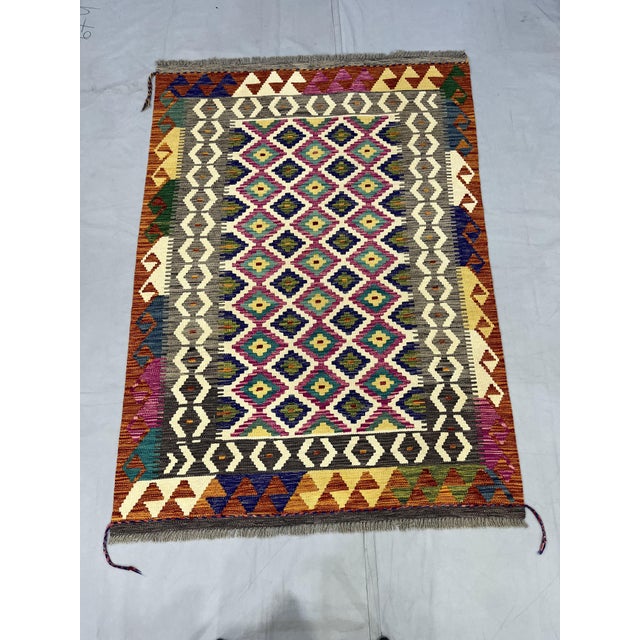 This Afghan Chobi flatweave kilim features a cream field with repeating colorful geometric motifs in rust orange,...