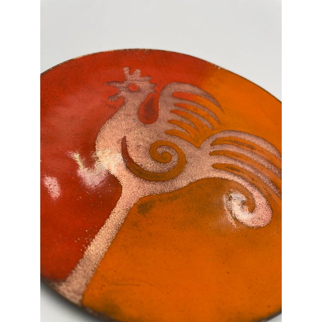 This modern yet whimsical trinket tray features a copper rooster motif. The image is created by shades of red enameling...