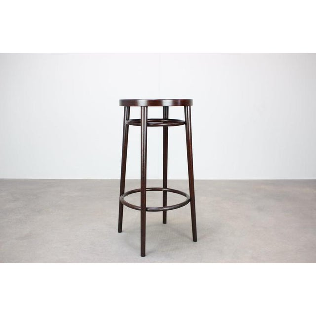 The stool features a round solid wooden seat supported by four slender legs with two circular bentwood reinforcing rings....
