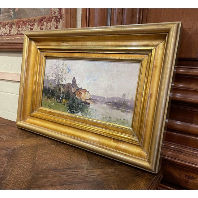 Painted in France circa 1890, this refined antique oil on canvas depicts a tranquil riverside village with a church rising...