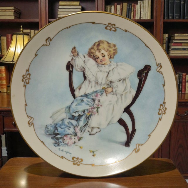 This vintage decorative plate features a charming design by Maud Humphrey Bogart titled "The Seamstress," perfect for...