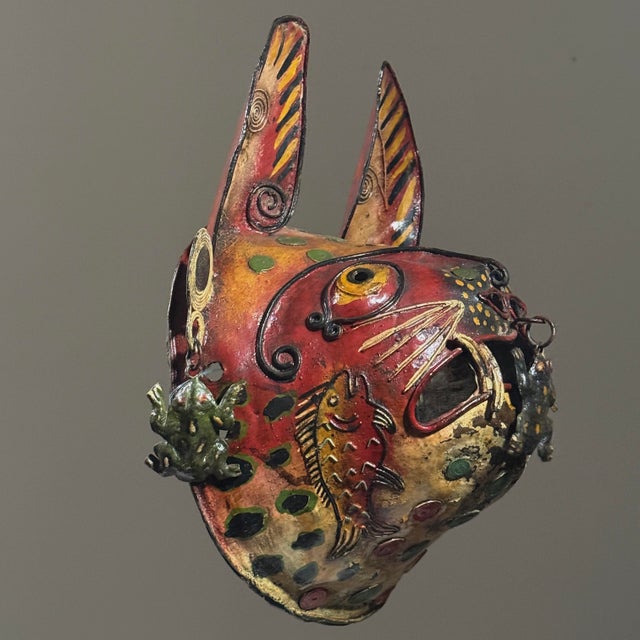 Crafted from polychrome metal, likely brass or copper, the mask represents a feline hybrid, its surface covered with hand...