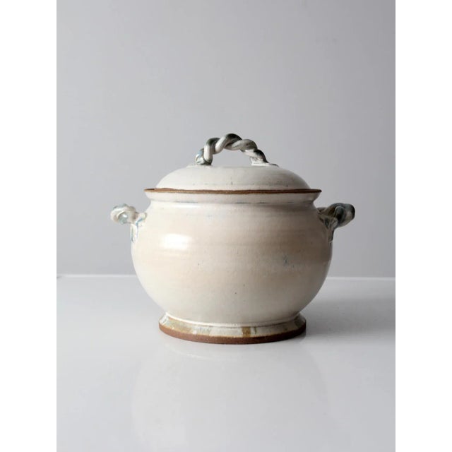 This is a vintage studio pottery tureen. The hand-crafted covered bowl features a white glaze with blue twisted handles on...