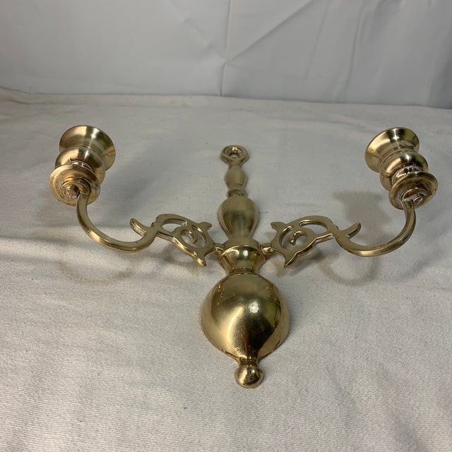 A vintage solid brass double-arm wall-mounted candle sconce featuring an ornate scroll design and a central hanging loop....