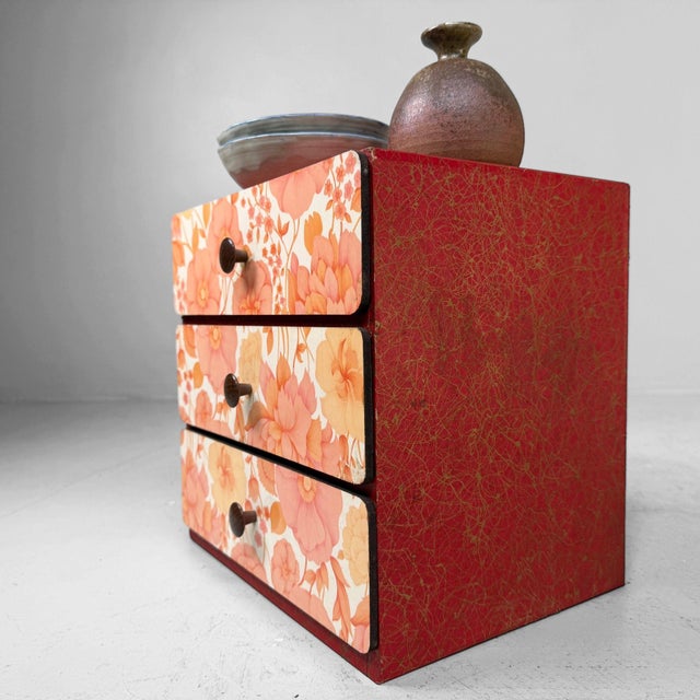 A charming Japanese tabletop drawer cabinet with three drawers, decorative floral fronts and a warm red-and-gold finish....