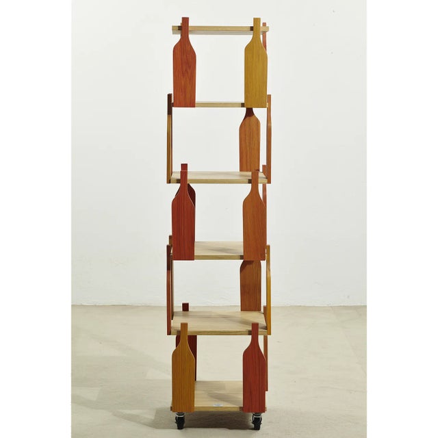 Wooden storage unit with casters. The intermediate shelves are connected by silhouettes of wooden bottles in various warm...