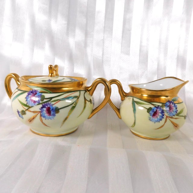 This is a cream and gold sugar bowl with lid and matching creamer. They have purple and blue flowers and greenery all...