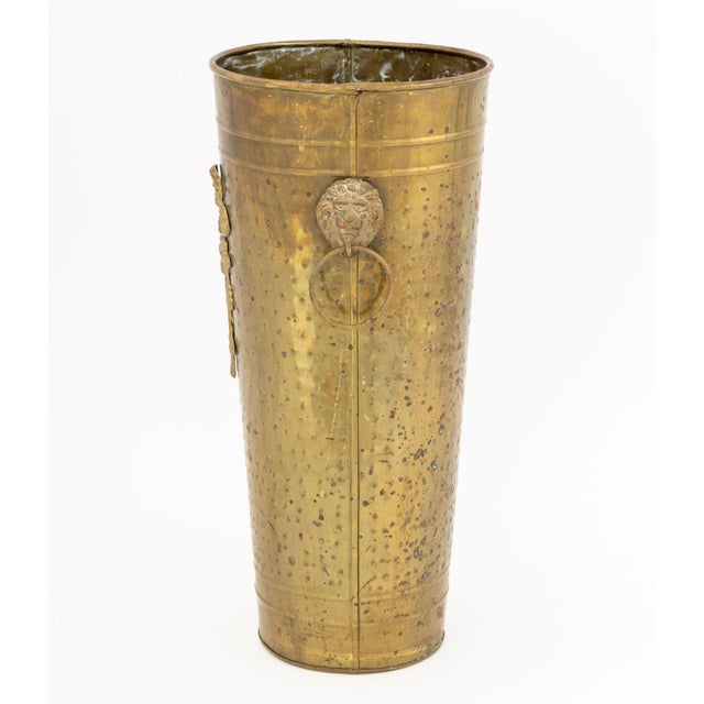 Arts and Crafts Hammered Brass Umbrella Stand, circa 1900, with two lion masque ring handles and ribbon foliate applique....