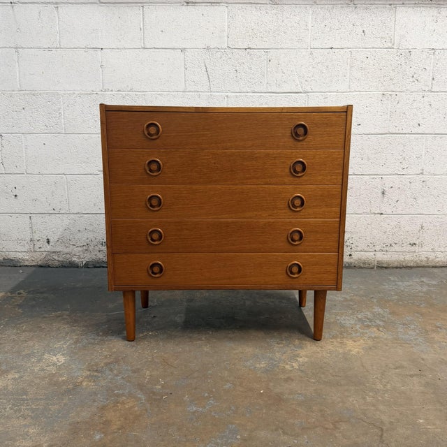 A compact Danish teak chest from the 1960s with five wide drawers and sculpted circular pulls. The repeated rings bring a...