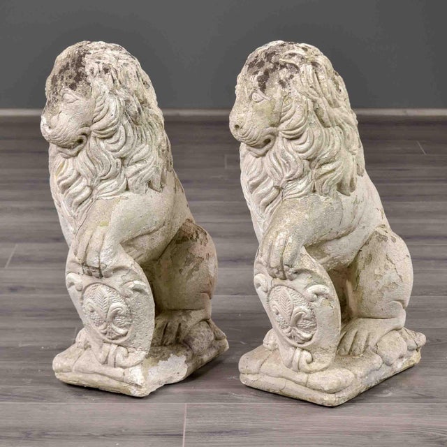 This matched pair of cast stone garden lions depicts each figure seated with one forepaw resting on a shield bearing a...