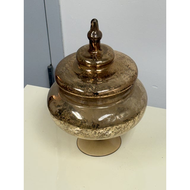 Design Plus Gallery presents an Antiqued Mercury Glass Footed Lidded Jar. The rounded glass vessel rests upon a circular...