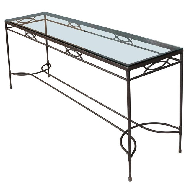 A console table crafted from iron with a dark patinated finish, topped with a glass surface. The clean, linear frame is...